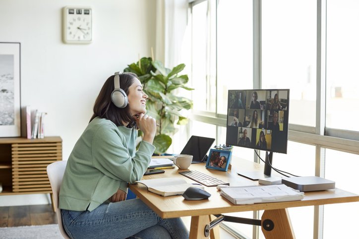 Smiling businesswoman discussing with colleagues through video call. Female professional is sitting in bright home office. She is listening through headphones.