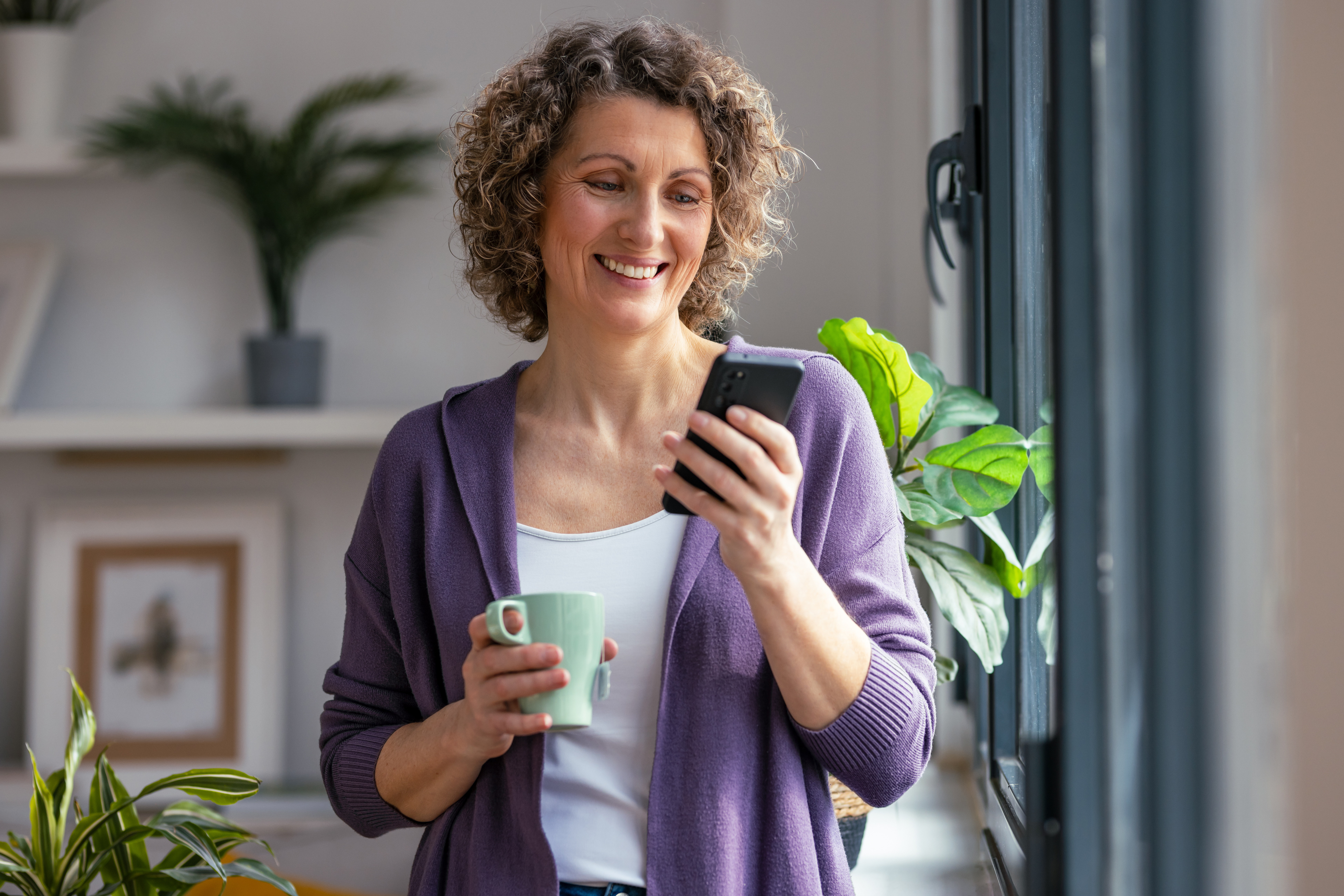 Shot of pretty mature woman using her mobile phone while drinking coffee near to the window in the living room at home