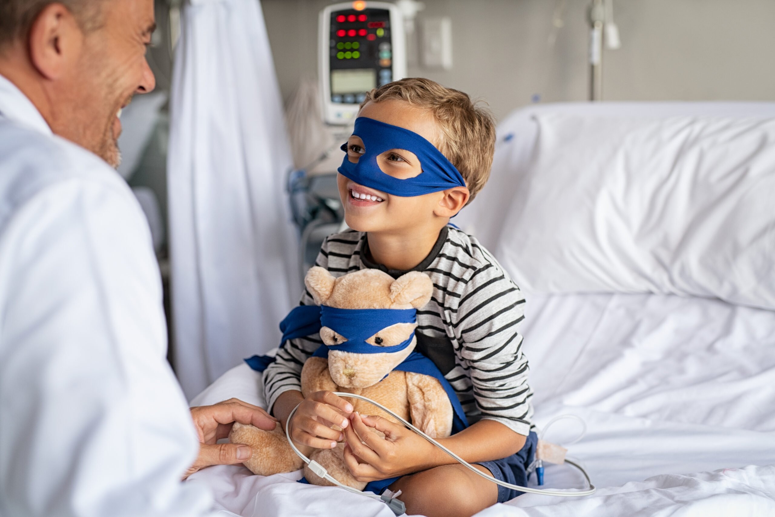 Cute little boy with superhero mask embracing teddy bear while playing with doctor and sitting on hospital bed. Playful child dressed as superhero holding stuff toy talking to pediatrician. Mature doctor visiting his little patient in hospital.